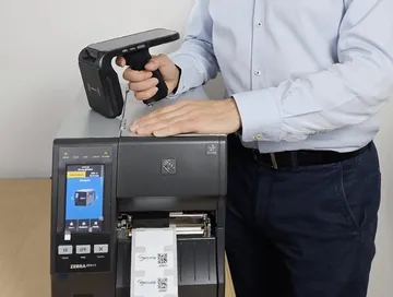 Ręczny czytnik RFID Chainway C5 w dłoni, oparty o drukarkę etykiet RFID Zebra ZT411, z której wychodzą etykiety z kodem QR