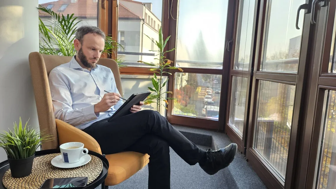 Piotr Szanser notuje rysikiem na tablecie, siedząc w fotelu przy oknie: spisywanie wymagań przed wyborem systemu